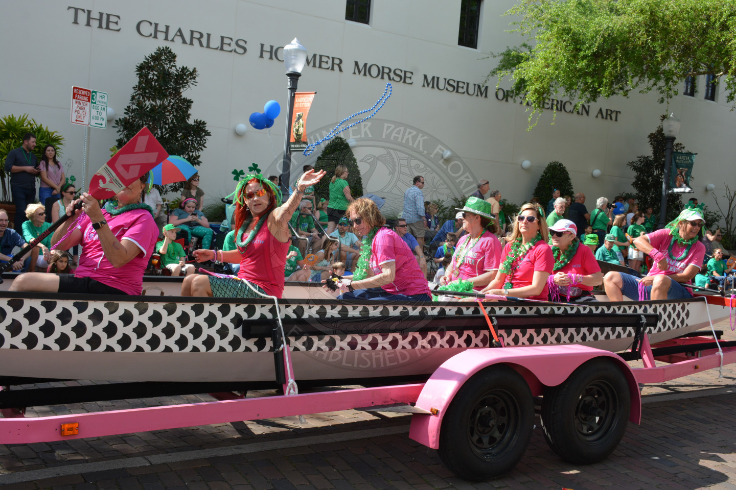 Approximately 10 people wearing similar shirts in a canoe or boat being pulled on a trailer bed down the parade route