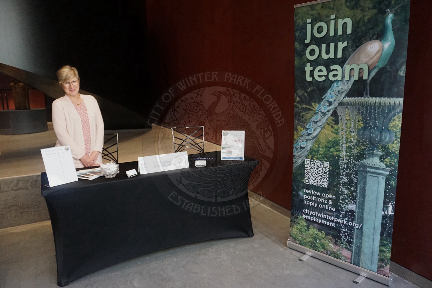woman standing posing for picture behind an 8-foot table next to a sign that states "join our team"