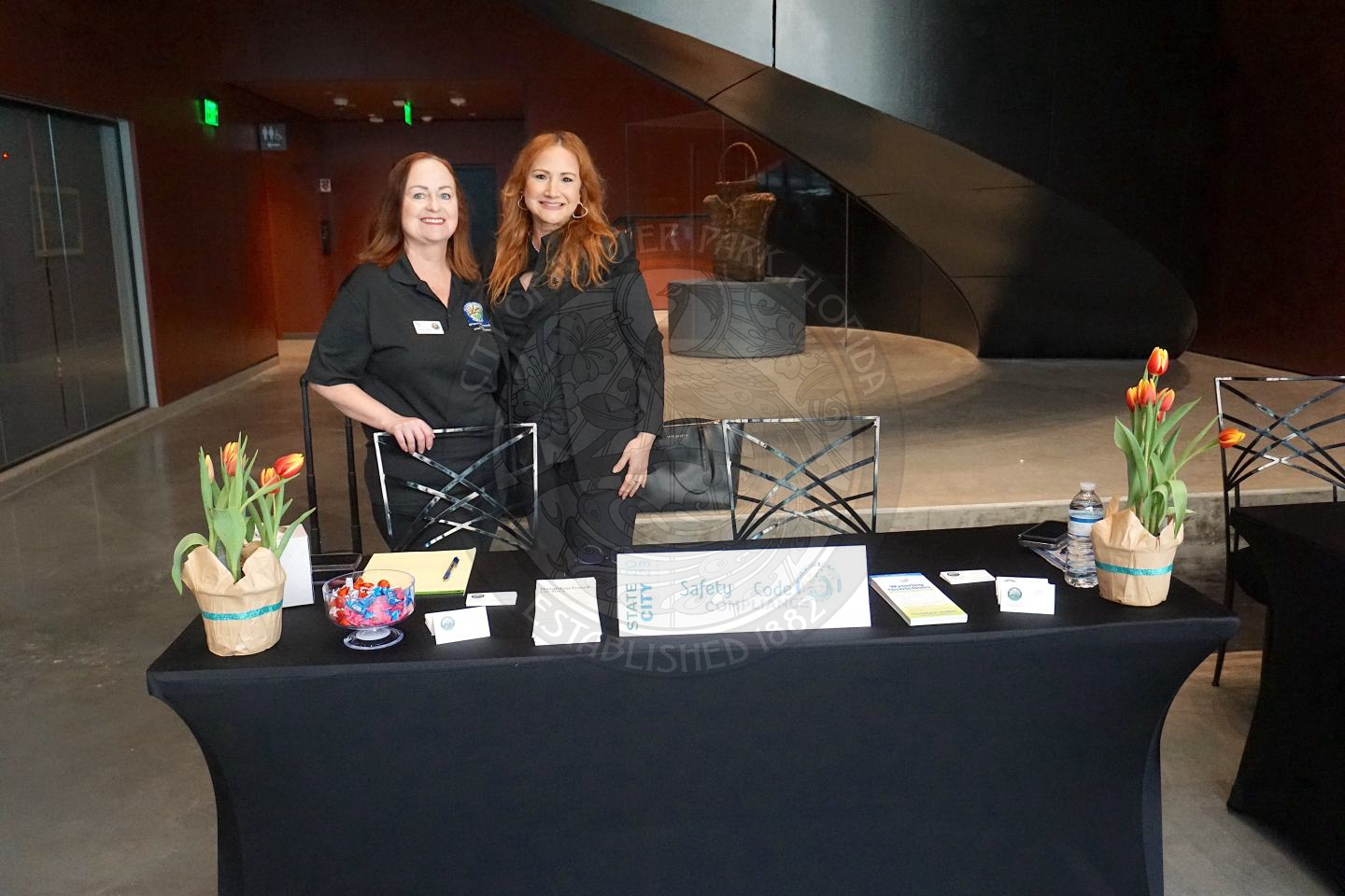 2 women standing behind an 8-foot table with a black tablecloth and paper with 2 flower pots on each side of the table.