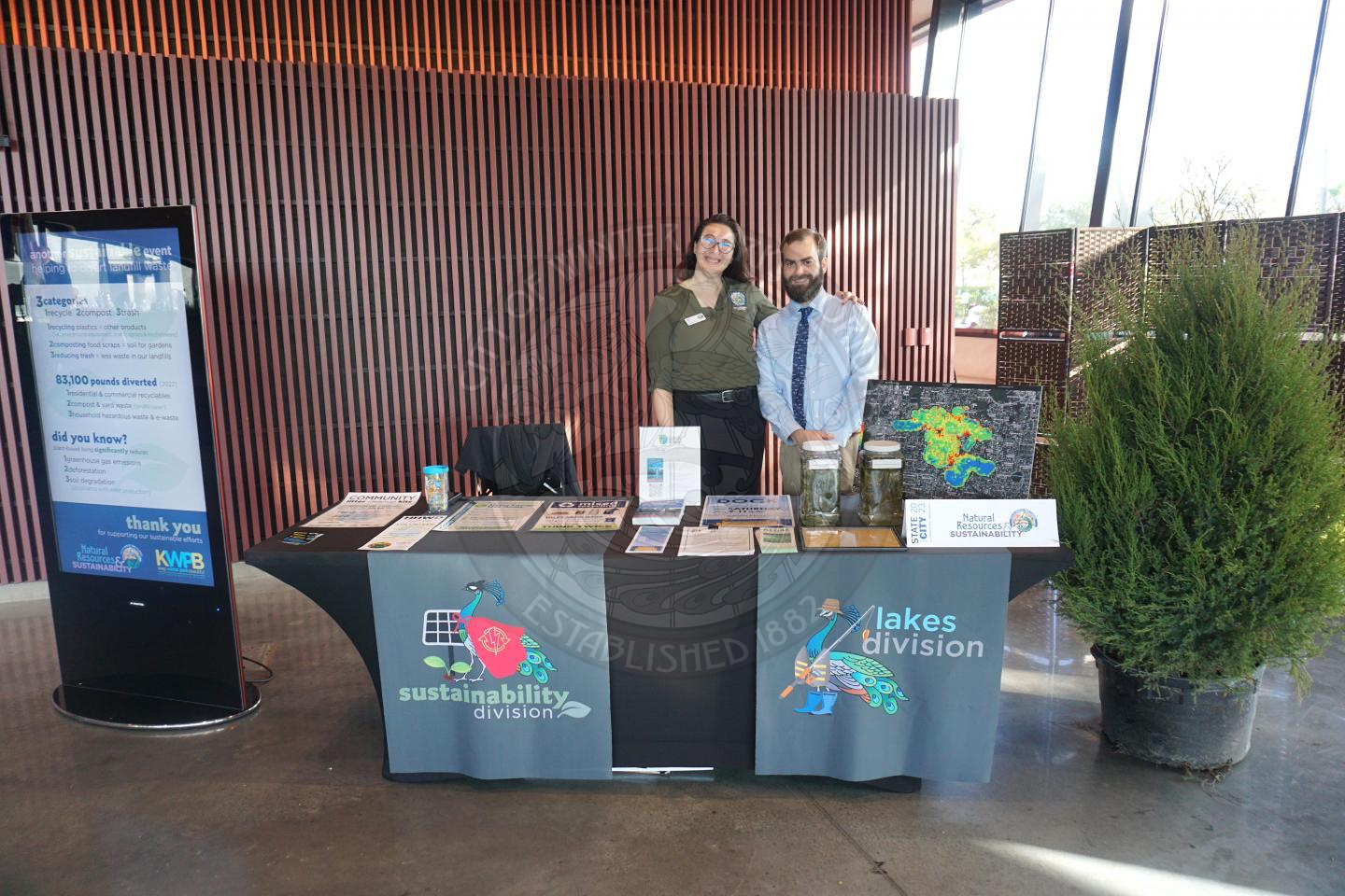 a man and woman standing behind a table with table runners that say Sustainbility Divison and Lakes Division.