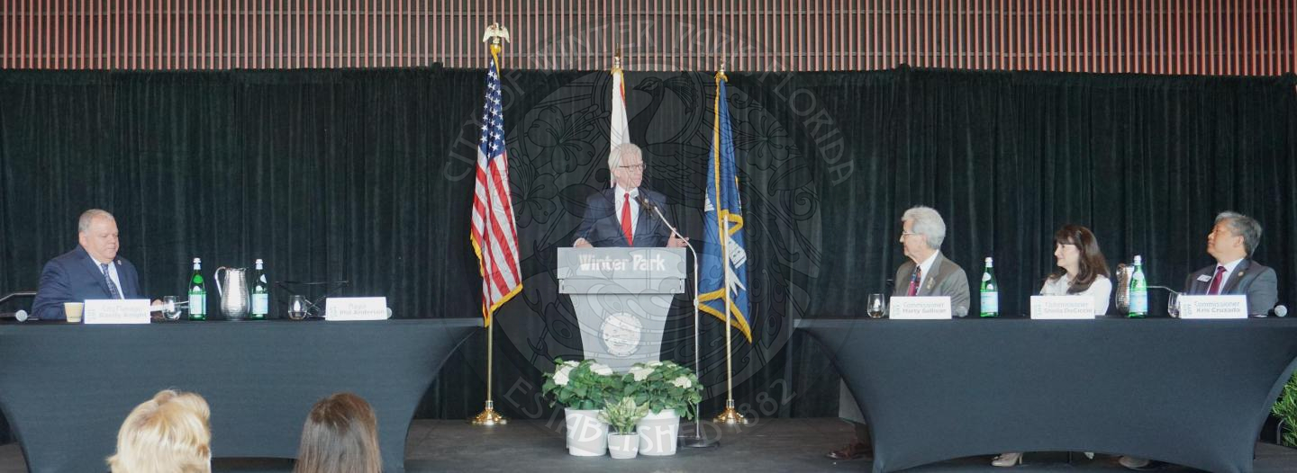 landscape shot of the stage with the mayor standing behind podium talking with a table on the left side of the podium with city manager sitting down. To the right of the podium with 3 other commissioners sitting down.