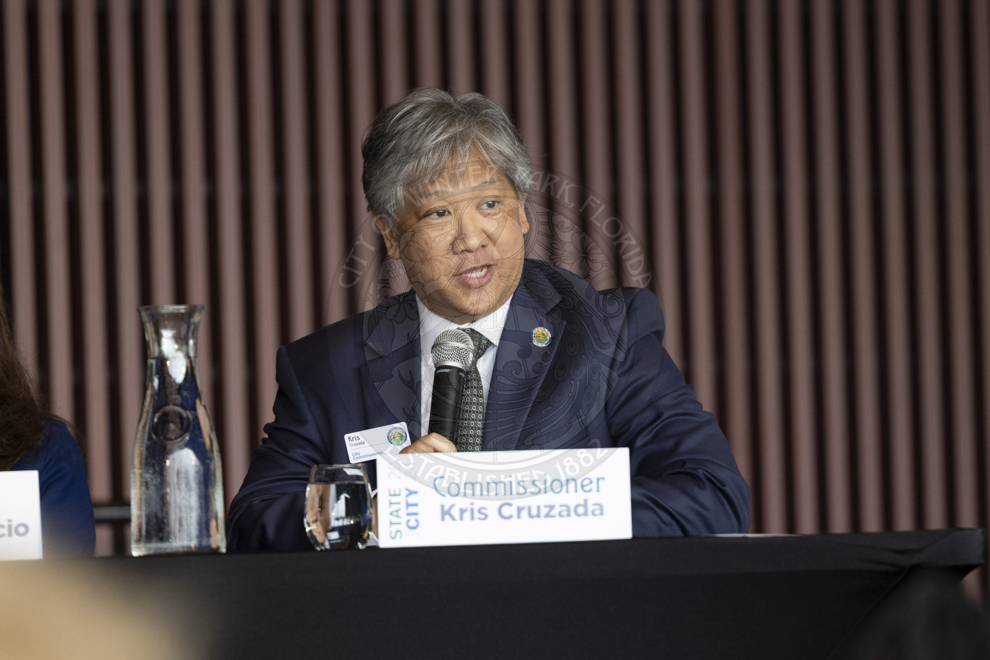 Commissioner Kris Cruzada sitting down at a table on the stage talking into the microphone speaking to the audience. There is a glass of water in front of him.