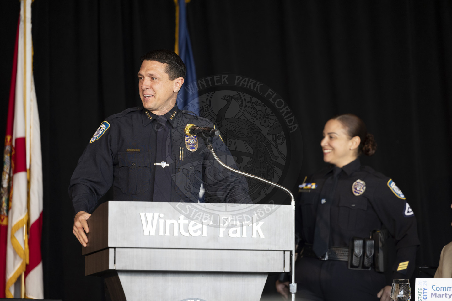Chief of Police stading behind the podium on the stage talking to the audience introducing the officer of the year who is standing behind him and to the right. 