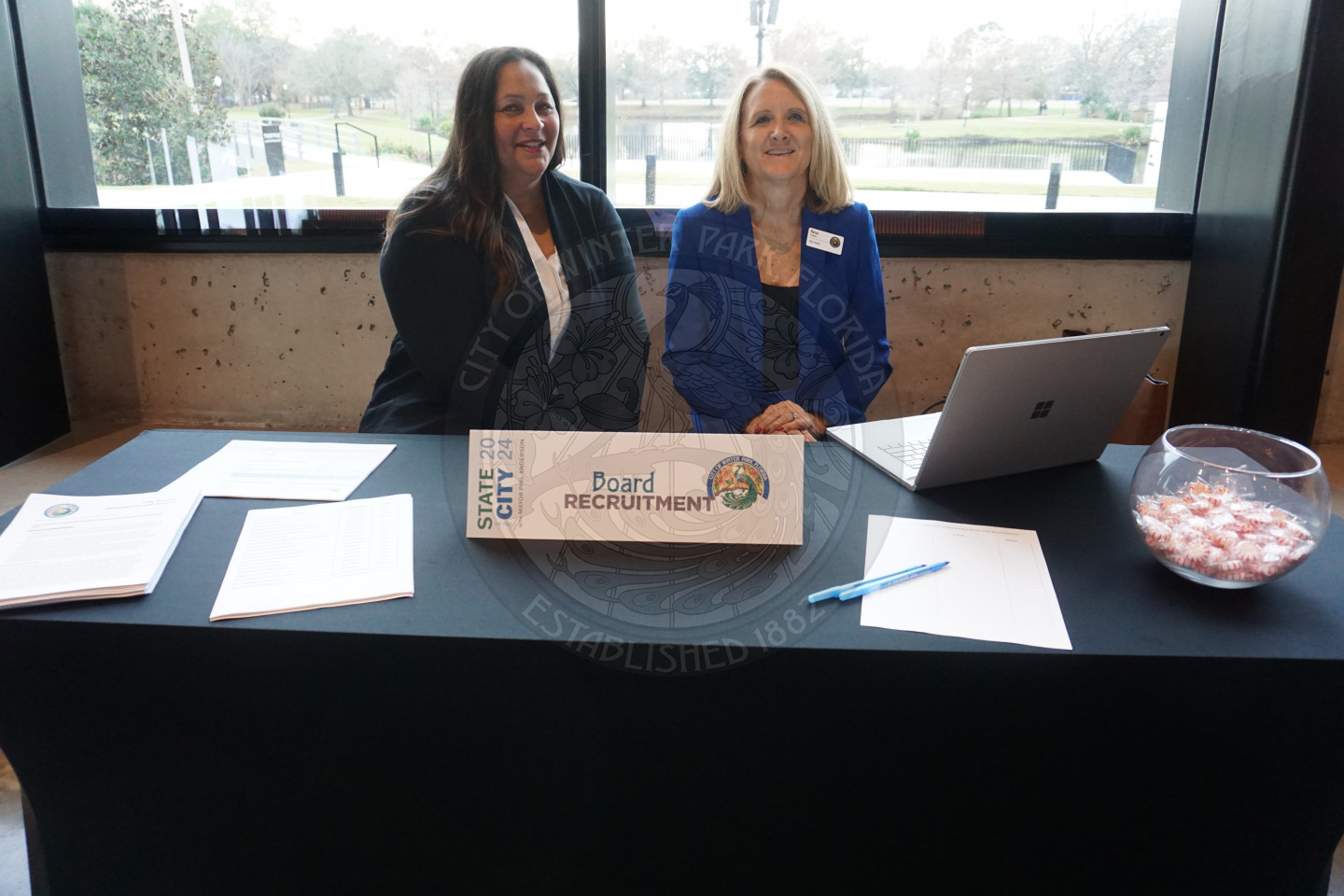 Two women sitting a behind a table that has paper, a table tent that says board recuritment, and a glass bowl of mints on the right side. There are two women sitting next to each other behind the table.