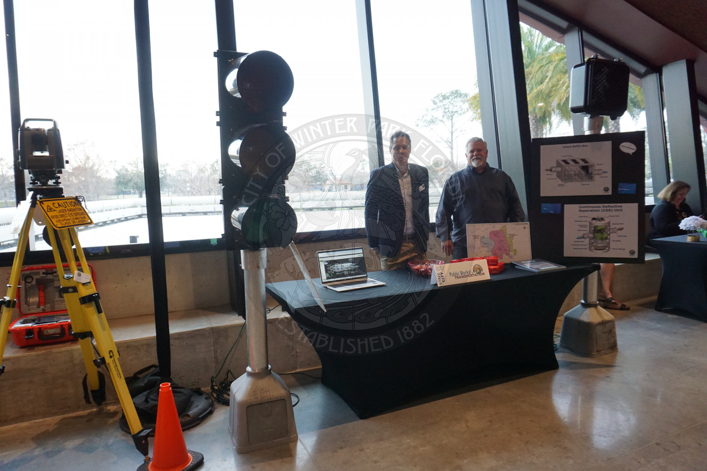 two men standing behind a table with a laptop and a table tent sitting on it. To the left of the table is traffic light that is about 10 ft tall. to the right of the table is a crosswalk sign.