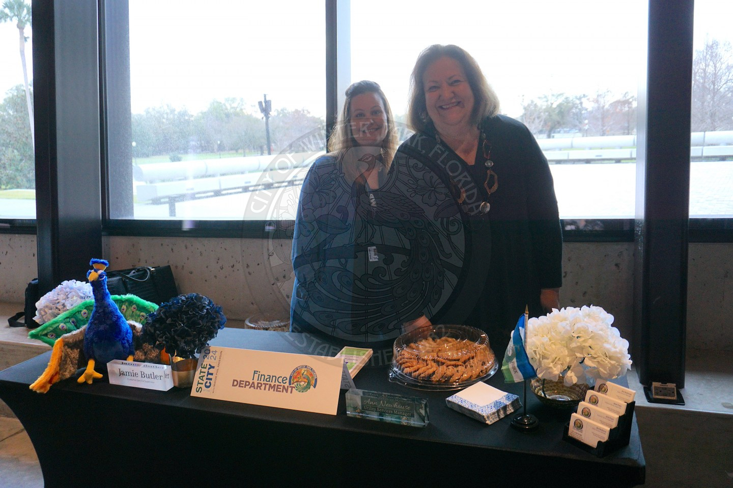 Two women standing next to each other behind a table that has some cookies, business cards, table tent that says "Finance Department", and a plush peacock.