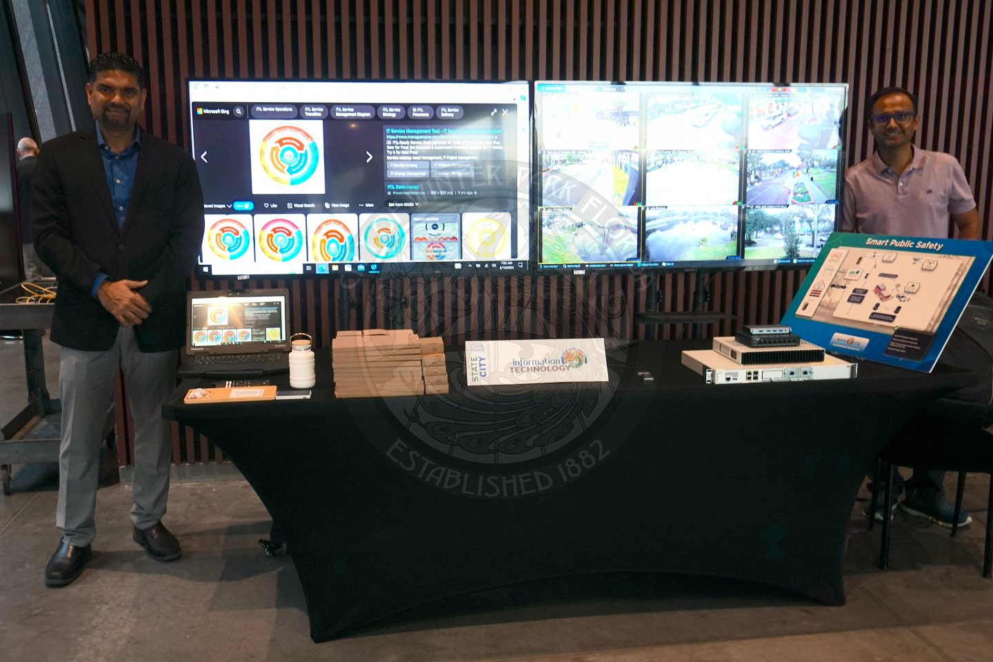 There is a table that is displaying information about the IT department. There are two TV's behind the table with two men standing next to a tv on each side. 