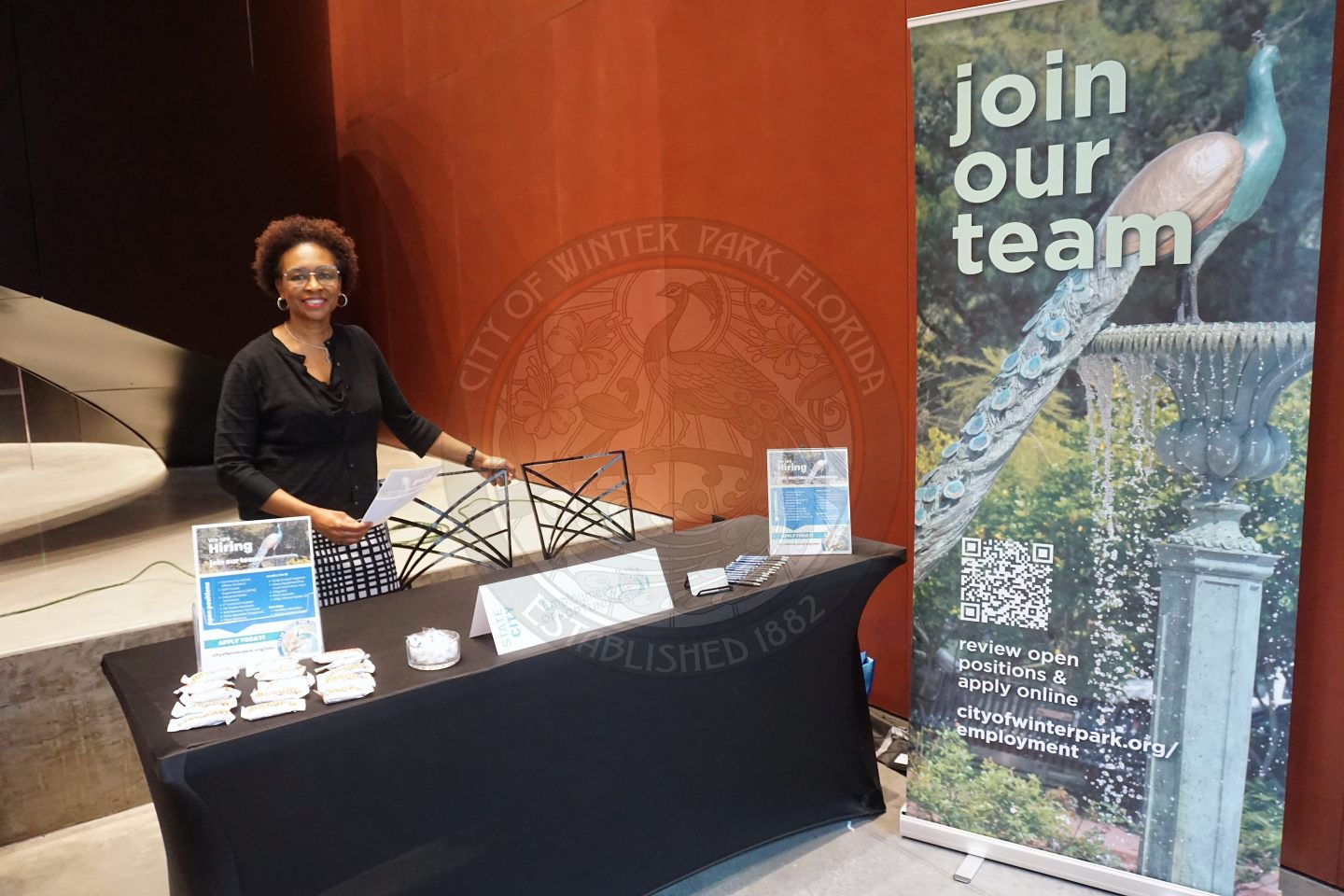 A woman standing behind a table smiling as she is pormoting to work for the City of Winter Park. There is a sign that is about 8 feet tall and says "join our team".
