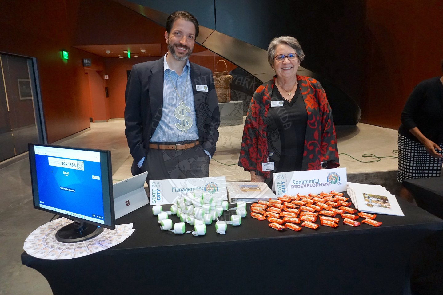 A man and a woman standing behind a table smiling. On the table you will find hacky sacks, 100 grands and Winter Park 100 dollar bills. 