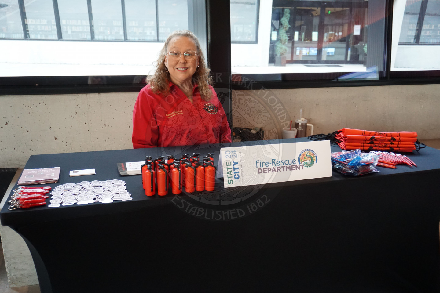 a woman smiling sitting behind a table with a table tent card that says "Fire-Rescue Department" with a bunch of promotional items around it.