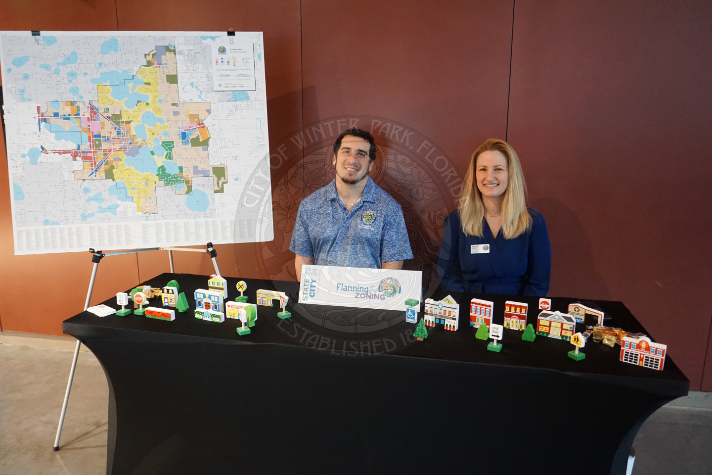 A man and woman sitting down behind a table with a block town. There is a posterboard next to the man with a GIS map of the City of Winter Park.