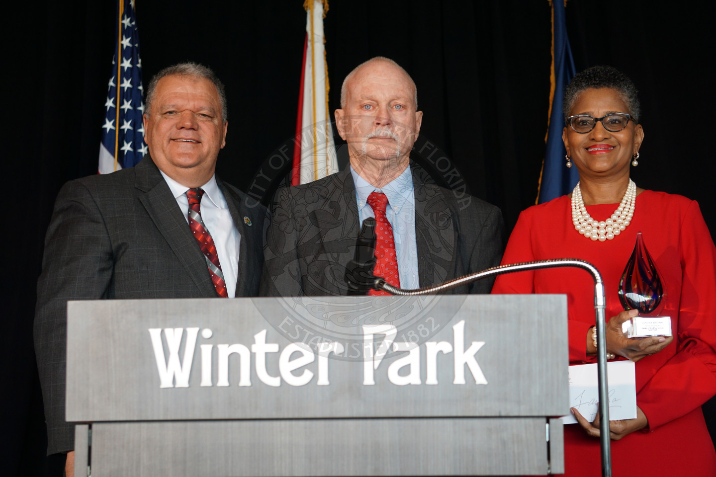 City manager Randy Knight, Director of Building &amp; Permitting Services Gary Hiatt and Employee of the year Sandra Brown who is holding an award standing behind a podium on a stage all smiling with joy.
