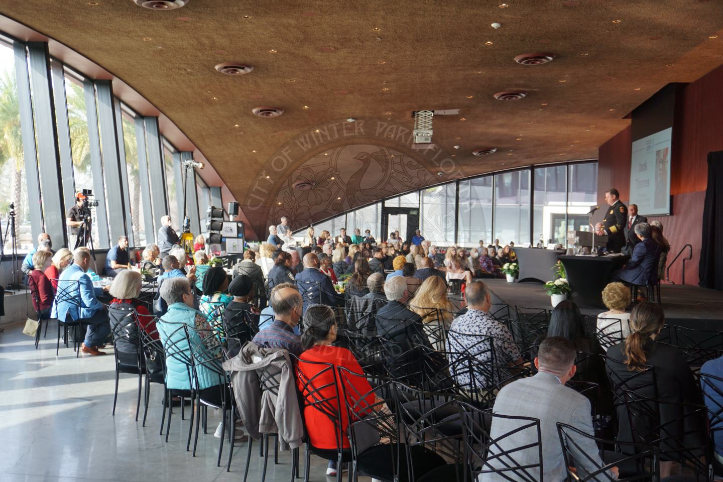 a ballroom filled with seats facing a stage with the winter park police chief talking to everyone seated in front of him. In the back of the room is a camera man with lights and camera recording the presentation.