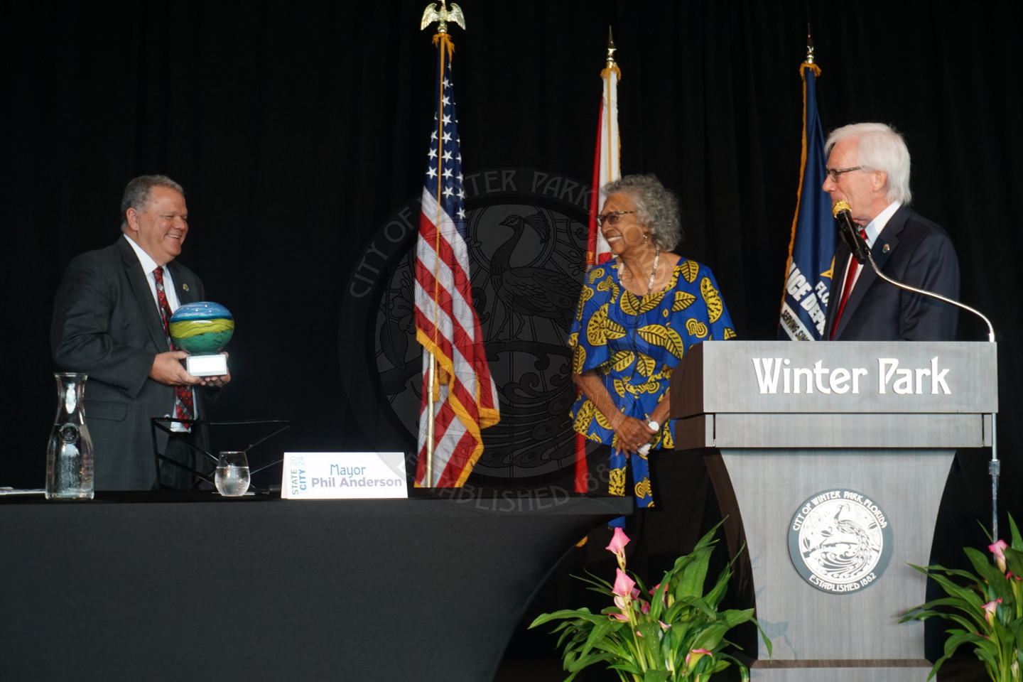 City manager Randy Knight holding the Mayor's Founder award standing behind a table walking toward Mary Daniels who is the recipient of the award. On the side is Mayor Phil Anderson standing behind a podium gleaming while watching Mary recieve her award.