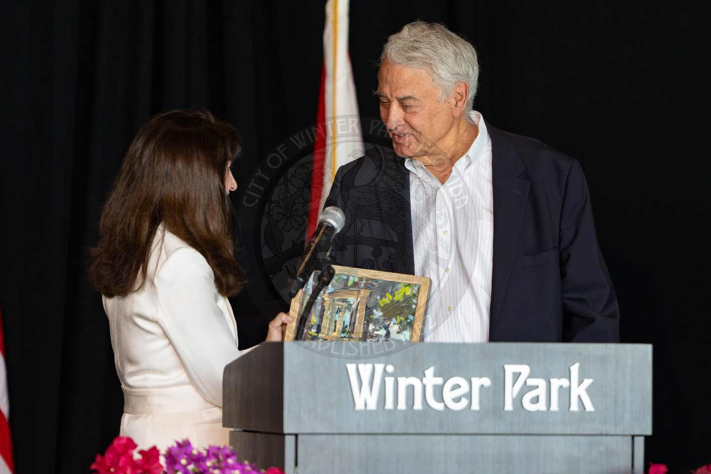 Mayor DeCiccio handing over a peice of art over to a man while standing behind a podium that says 'Winter Park'