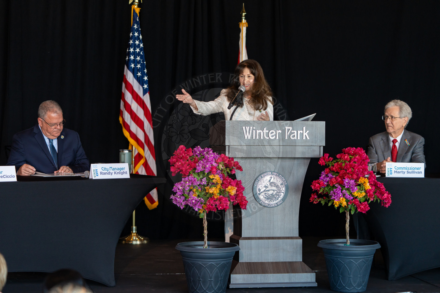 The female mayor of Winter Park standing behind a podium talking into a mircophone to guests on a stage with table next to the podium. 