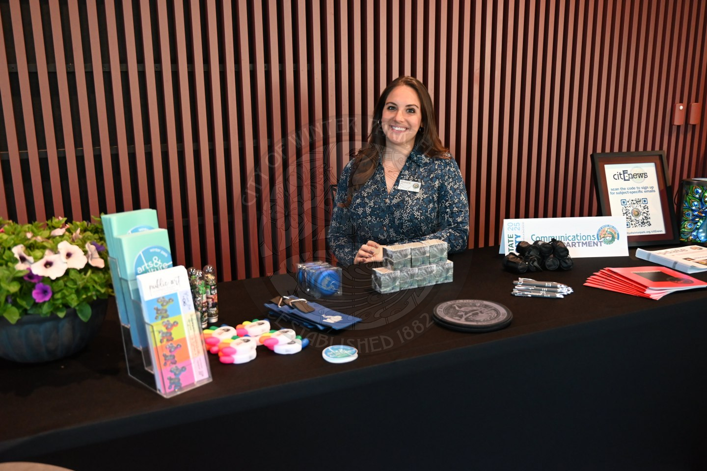 A woman sitting behind the desk of Communications table with promotional items.