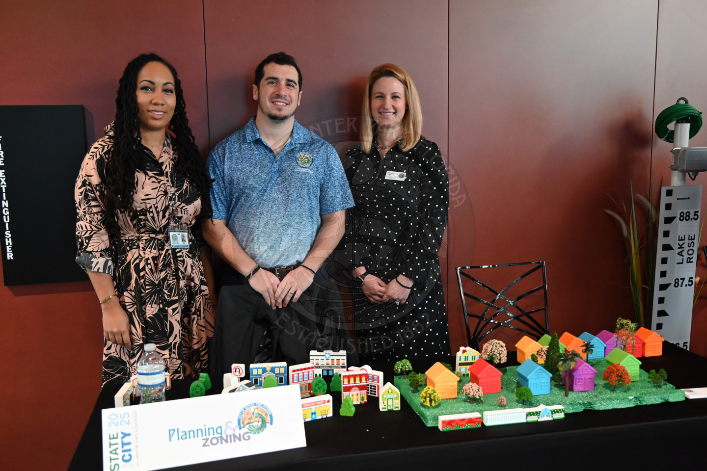 a man and two ladies standing behind a desk with a table full of buildings and houses. There is a sign that says "Planning &amp; Zoning" 
