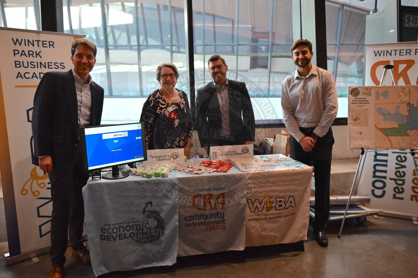 three men and a woman standing behind a table with banners about Winter Park Business Academy, Community Redevelopment Advisory, and Economic Development. 