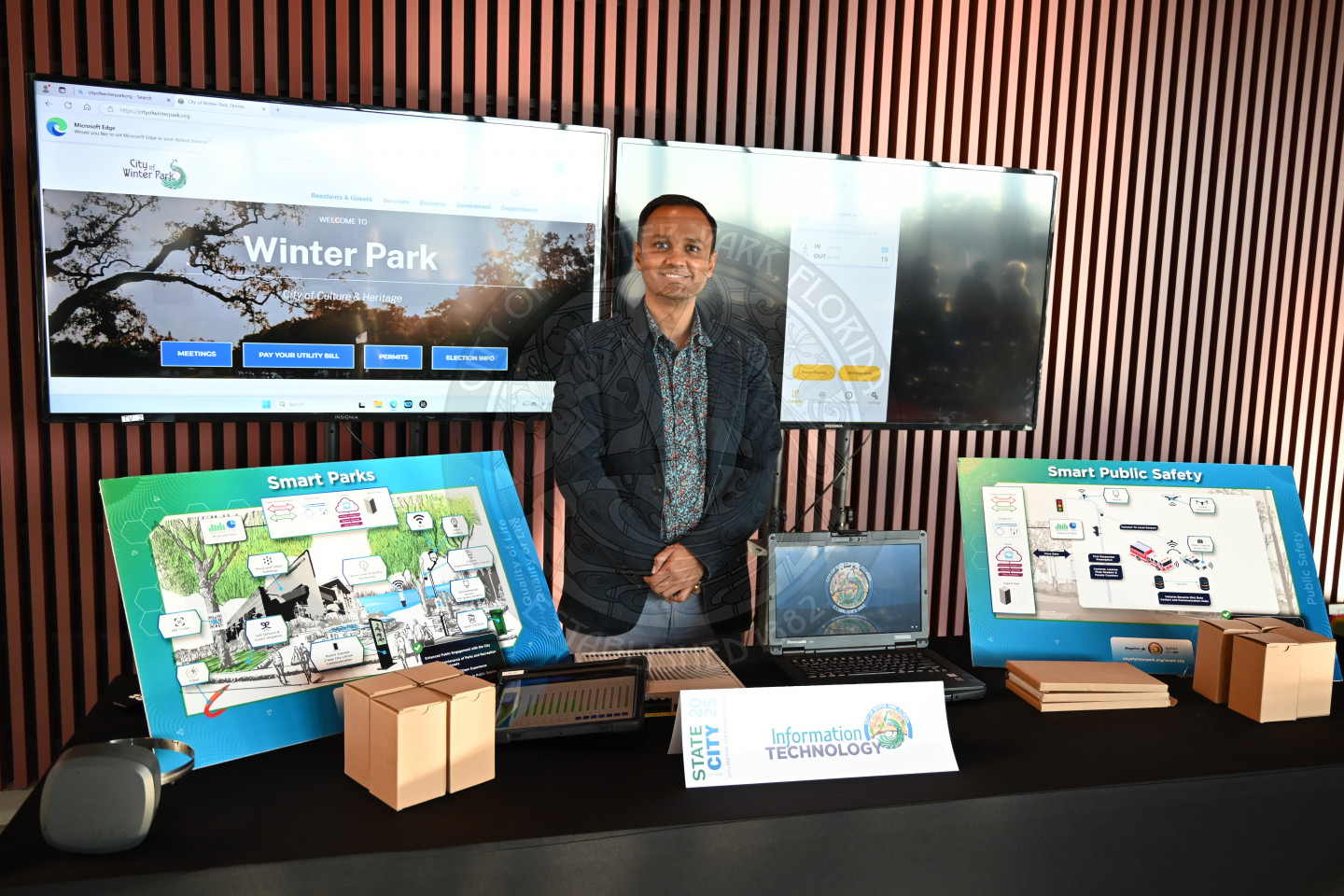 a man standing behind a table that has boards of what a smart city is and behind him are 2 tv's.