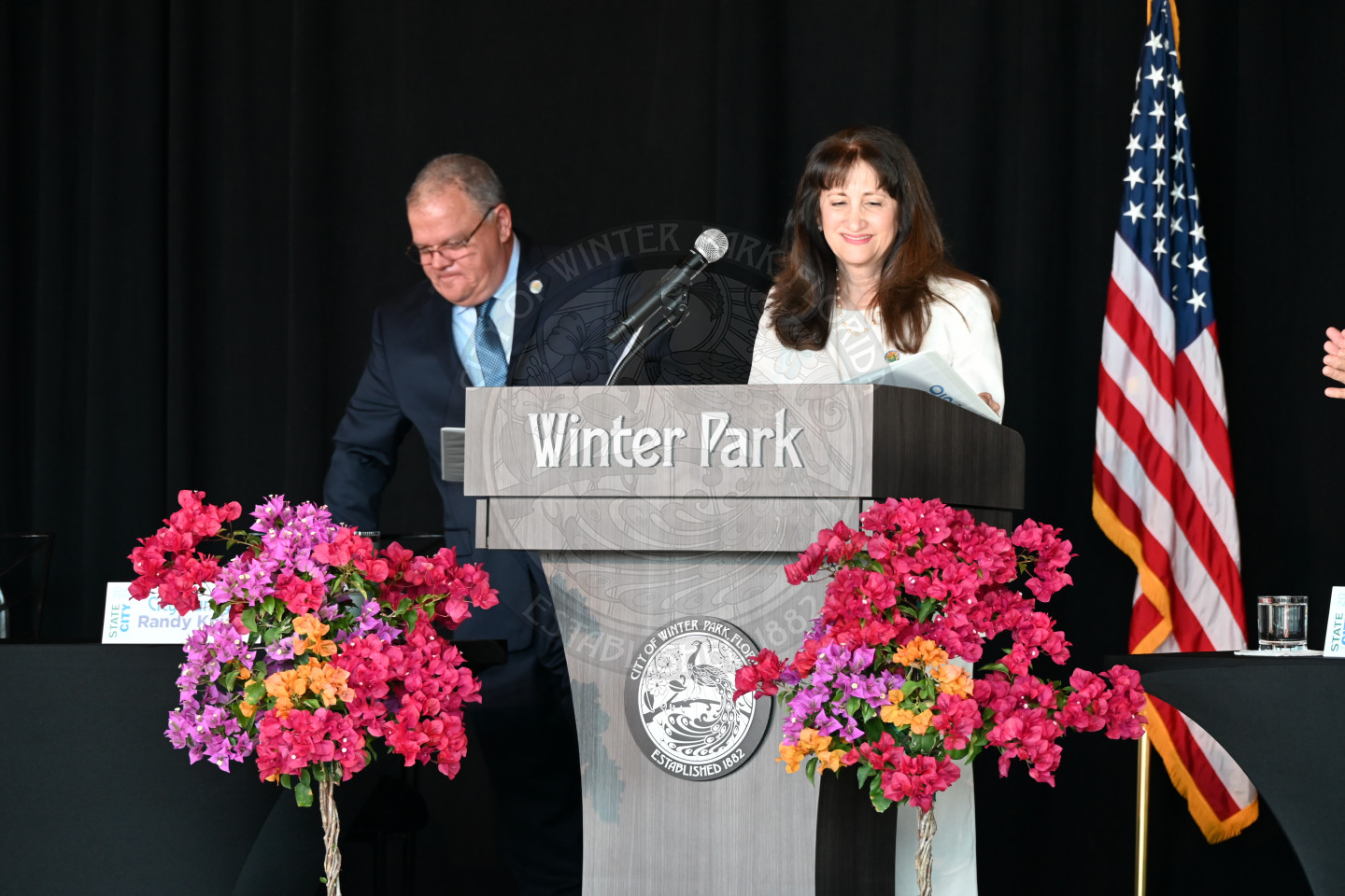 The female mayor standing on the stage behind a podium smiling with  a male standing behind her right side.