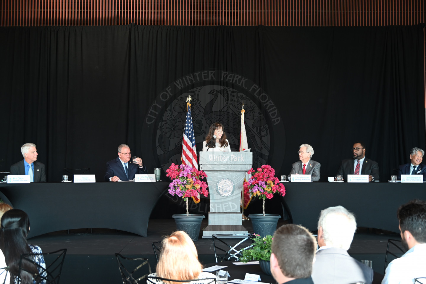 view of the stage with a female mayor standing behind the podium. Nex to the podium are tables on each side with men sitting at the tables.