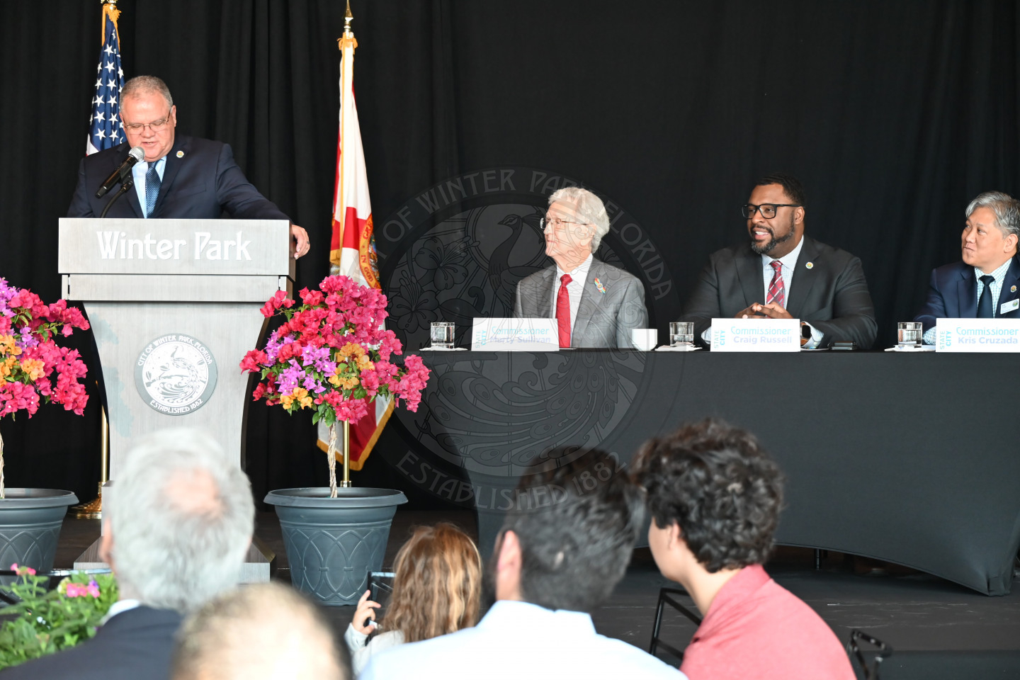 a male standing behind a podium talking to the microphone with a table seated with men sit to right of the podium.