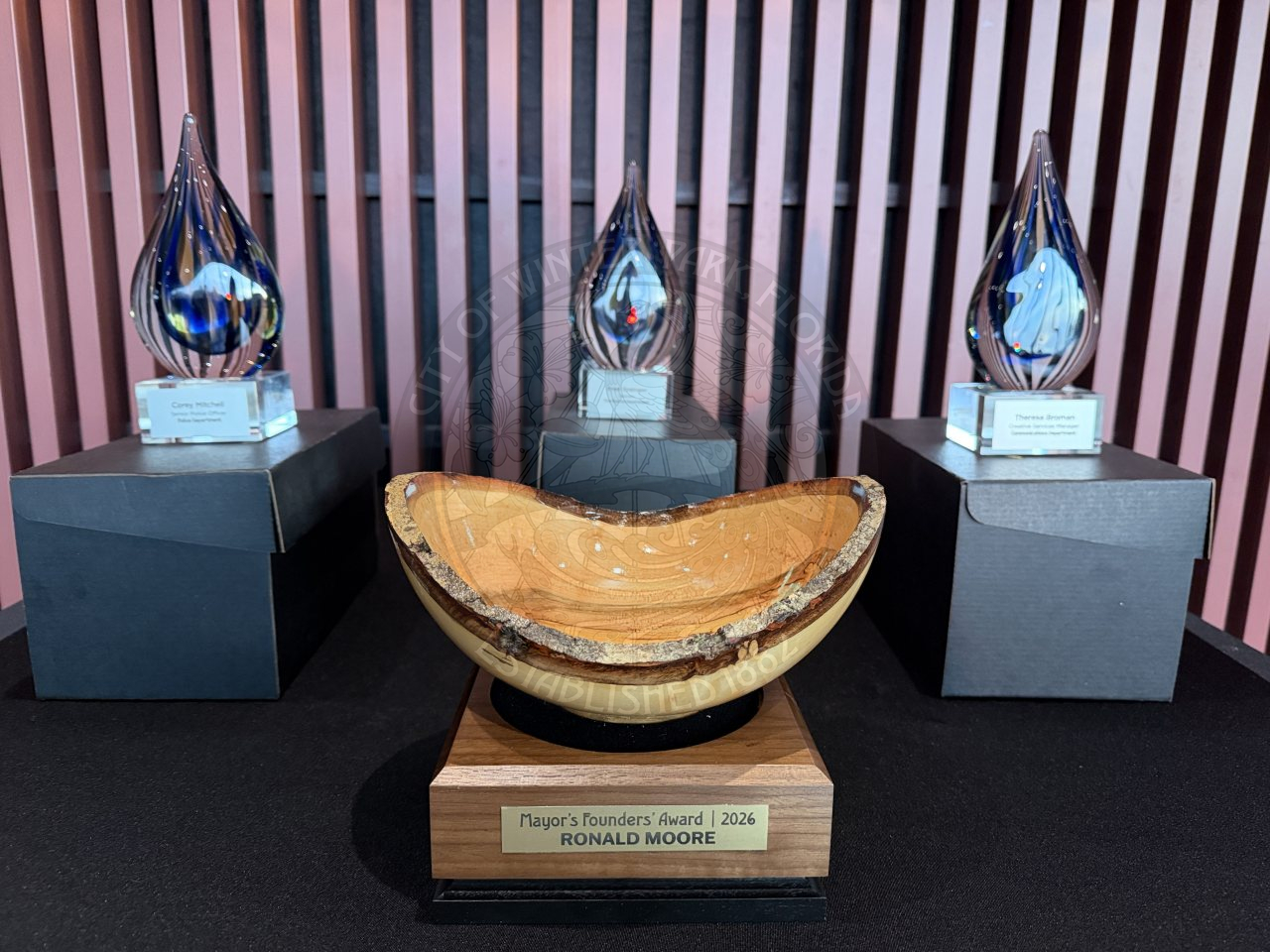 Three glass awards and a wooden trophy on display. The wooden trophy in front is engraved with “Mayor’s Founder’s Award | 2026 | Ronald Moore,” while the glass awards are set on top of black boxes behind it.