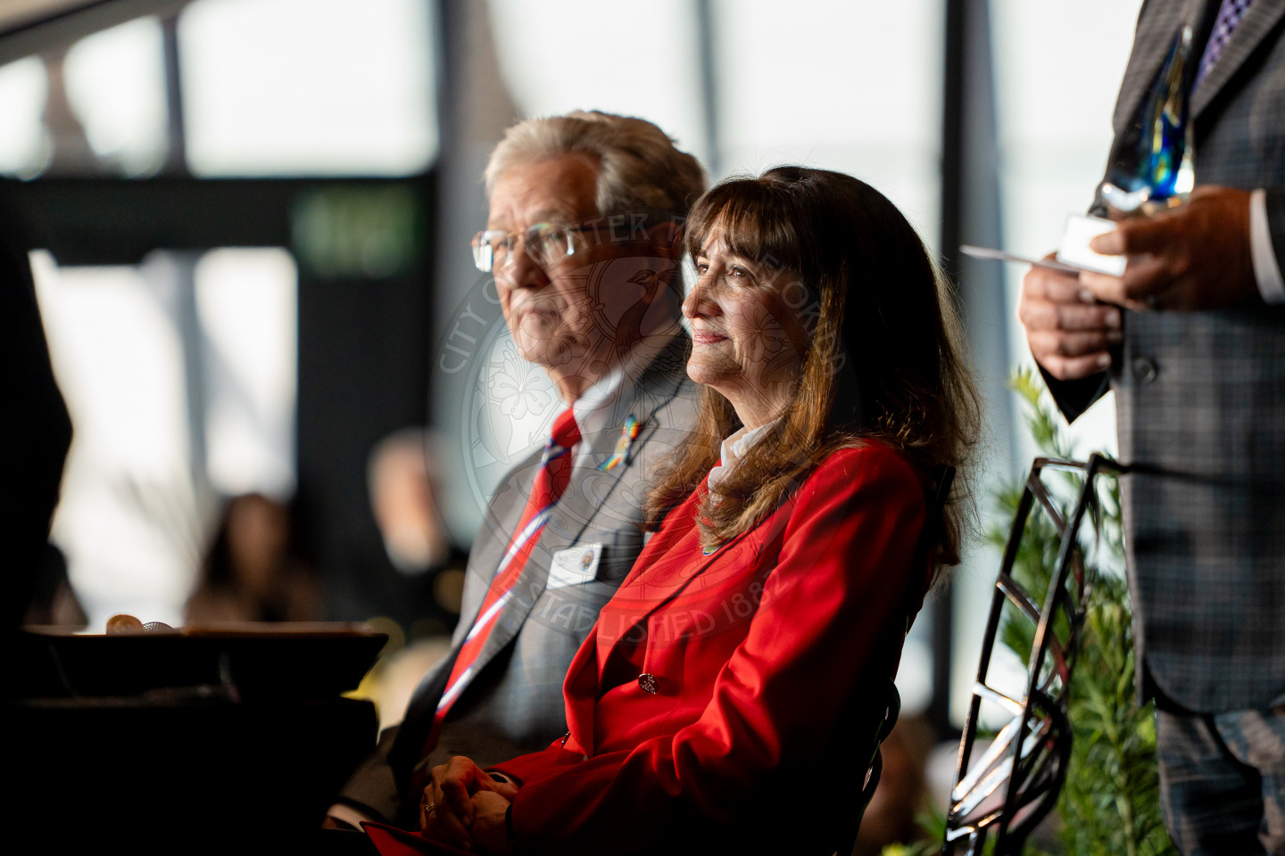 A man and a woman, both wearing business attire, sit side by side, attentively facing forward at an indoor event. The woman is in a red blazer, and the man is in a gray suit with a red tie. Other people are blurred in the background.