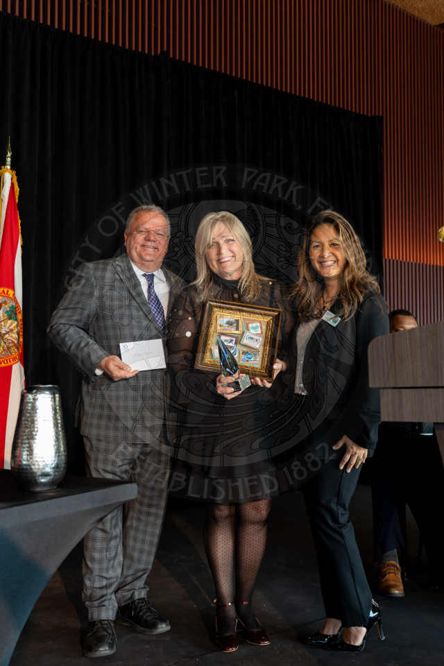 Three people stand together at an indoor event; the woman in the center holds a plaque and a glass award. The man on the left holds an envelope. They are smiling, dressed formally, and a Florida state flag is visible in the background.