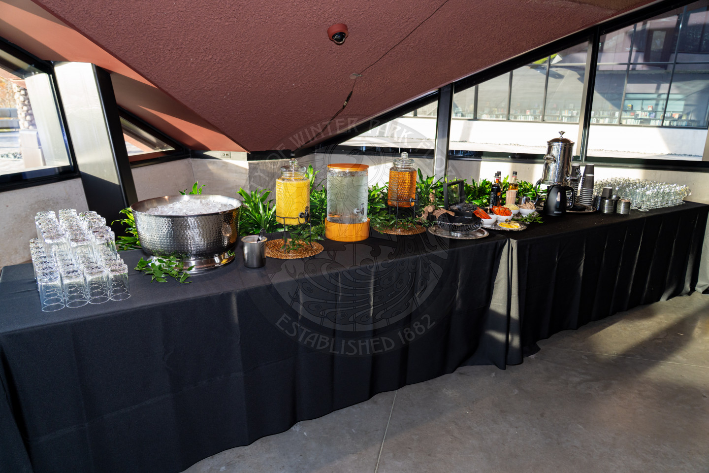 A long table covered with a black tablecloth holds drink dispensers with orange juice and iced tea, a large ice bucket, coffee urn, glassware, cups, and assorted condiments, set up near large windows.