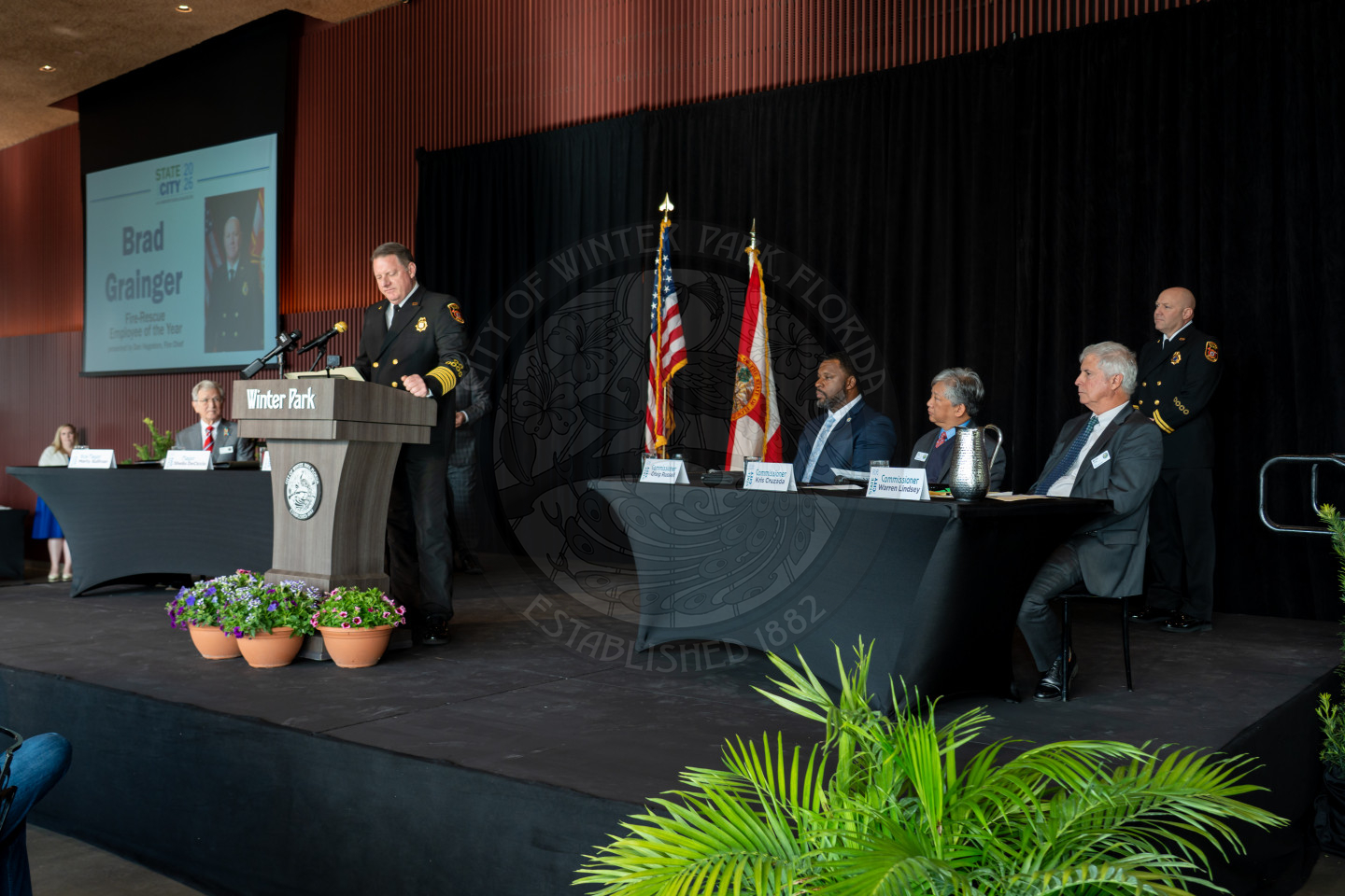 A police officer speaks at a podium labeled Winter Park during a formal indoor event with an audience, flags, and five seated panelists at a long table on stage. A screen to the left of the speaker displays Brad Grainger.