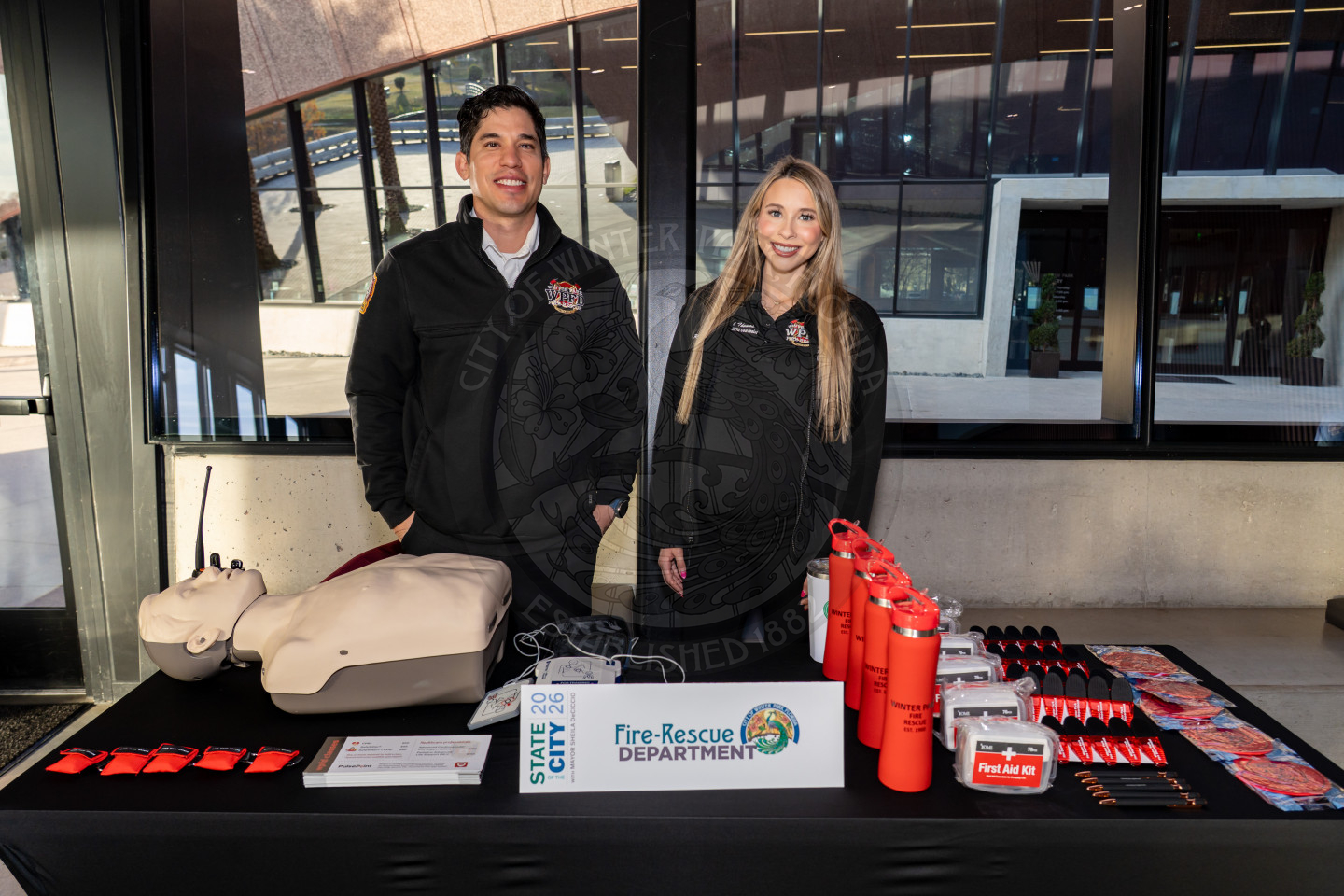 Two people in Fire-Rescue Department uniforms stand behind an information table with CPR mannequins, brochures, water bottles, and first aid kits at a public event indoors.