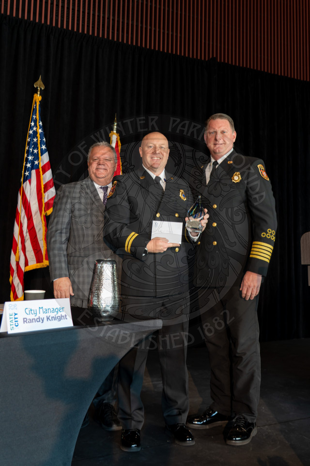 Three men stand together on a stage, smiling for a photo. Two are in formal fire department uniforms, one holds an award and envelope with an American flag behind the group.