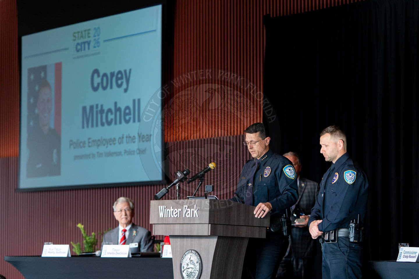 Two police officers stand at a podium labeled Winter Park, presenting an award. Behind them, a screen displays Corey Mitchell, Police Employee of the Year. Two other men sit at a table in the background.