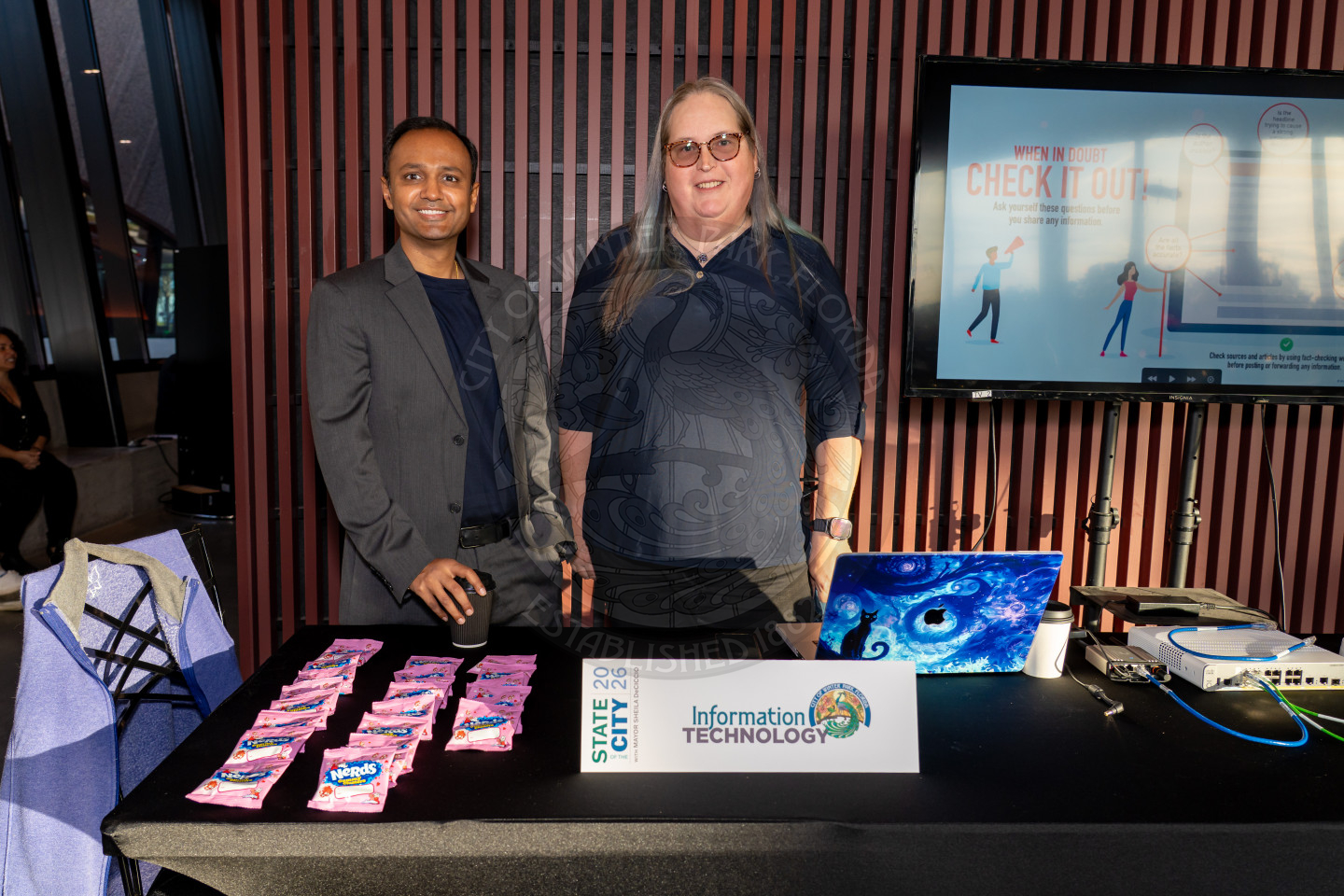 Two people stand behind a table with a laptop, candy, and an Information Technology sign. Behind them is a large screen displaying a presentation and tall vertical wall panels.