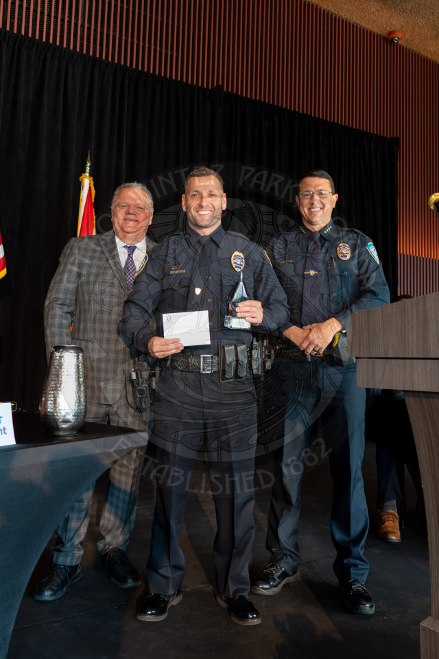 Three men stand smiling at an awards ceremony. The man in the center, wearing a police uniform, holds a plaque and an envelope. The other two men, in a suit and a police uniform, stand on either side of him.