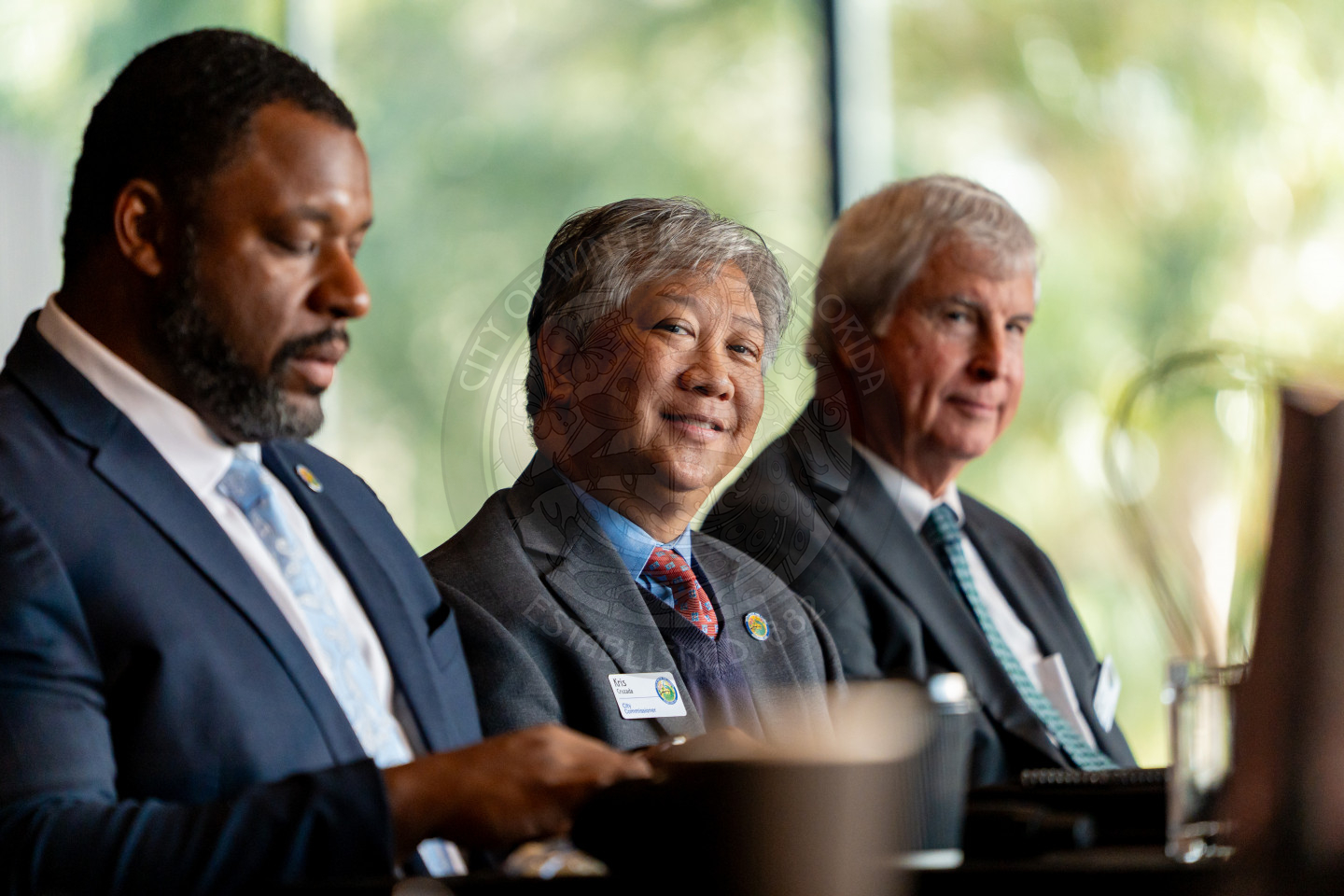 Three men in business attire sit side by side at a conference table, one smiling at the camera while the others look ahead or down, with blurred greenery visible through windows in the background.