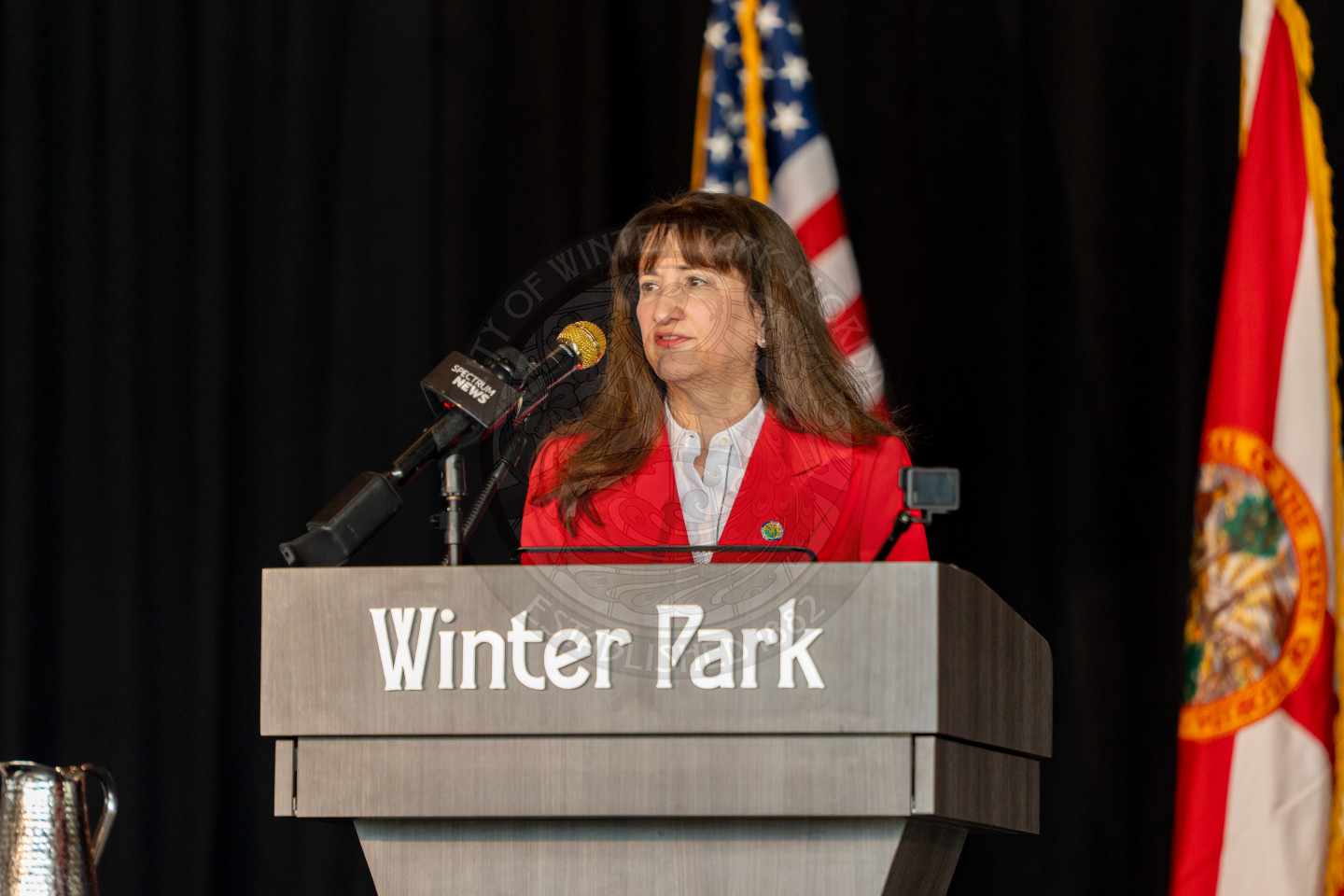 A woman in a red blazer speaks at a podium labeled Winter Park, with microphones in front of her and U.S. and Florida state flags in the background.