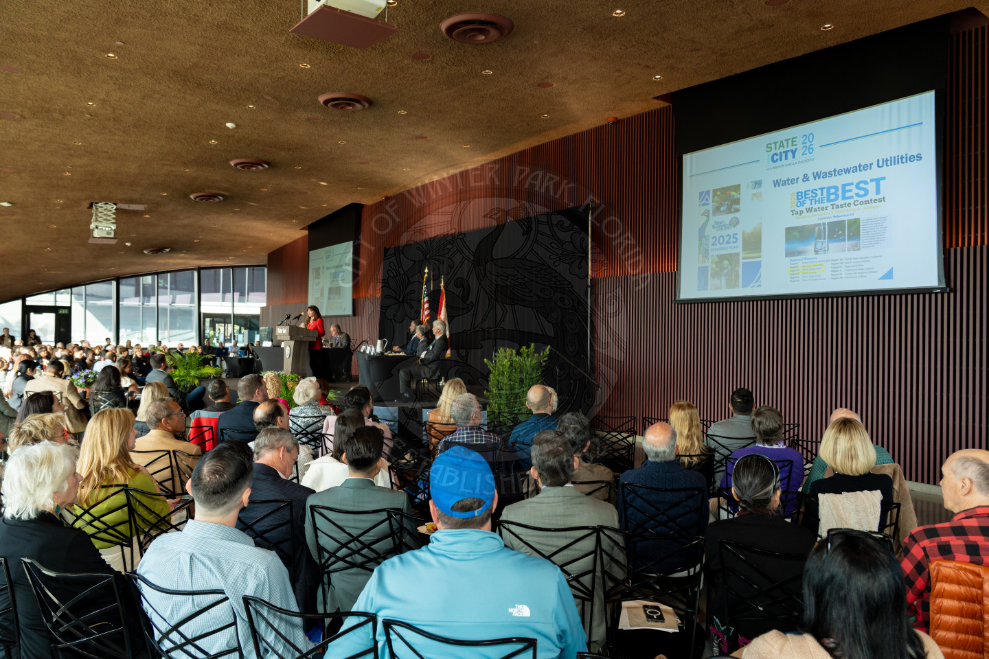 a woman speaking at a podium to a seated audience during a formal indoor event with floor to ceiling windows overlooking a park.