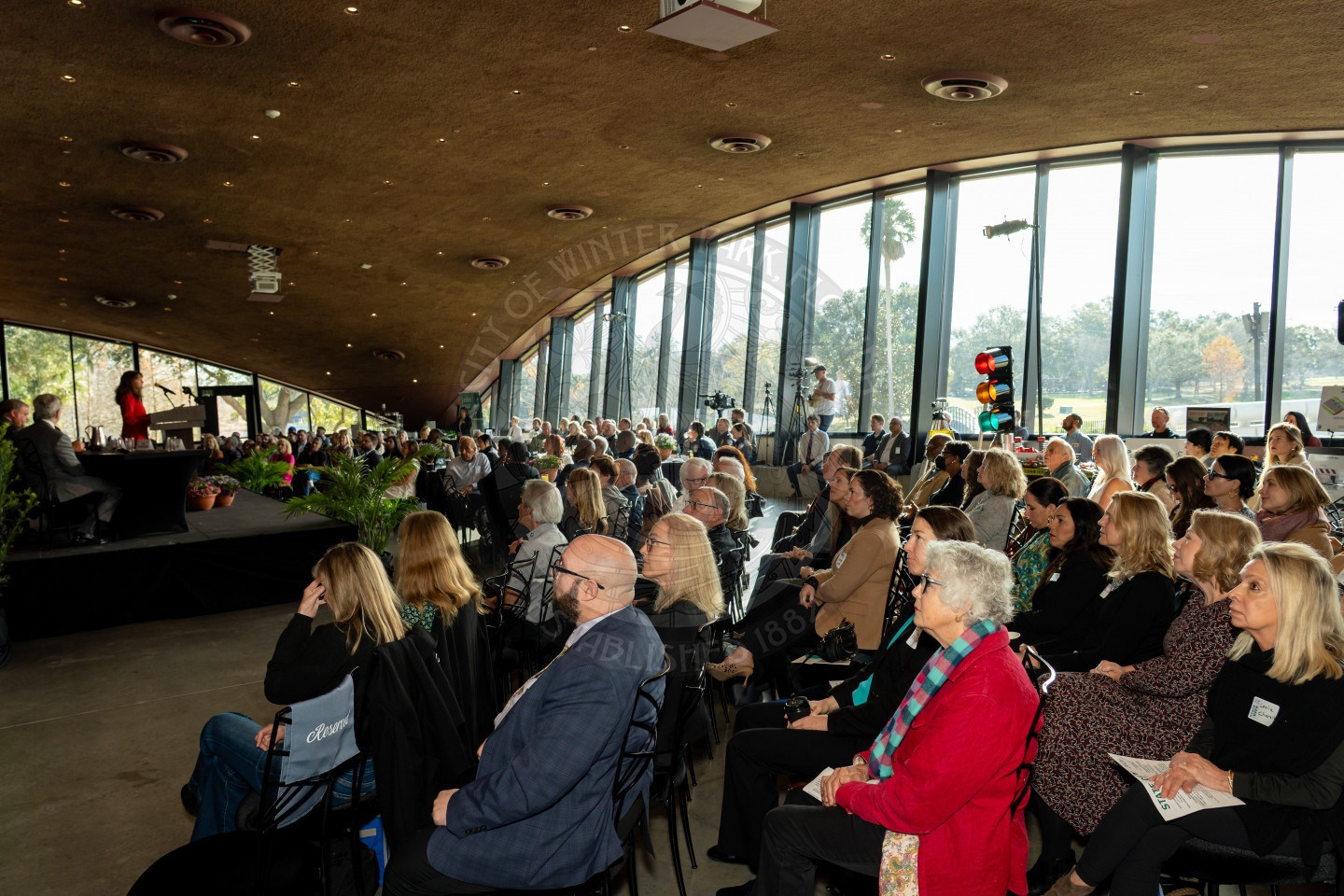 a woman speaking at a podium to a seated audience during a formal indoor event with floor to ceiling windows overlooking a park.