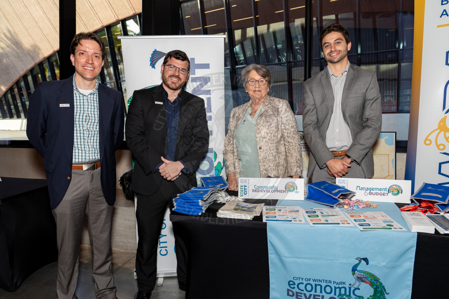 Four people stand and smile behind a display table with informational materials, brochures, and banners for the City of Winter Park Economic Development at an indoor event.