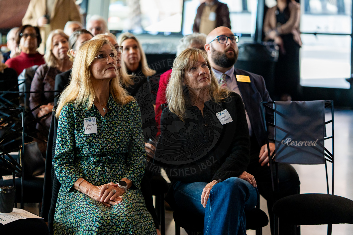 A group of people sit attentively at an indoor event. Two women in front, wearing name tags, face forward. Behind them, seated attendees listen, and one chair to the right in front is marked Reserved.
