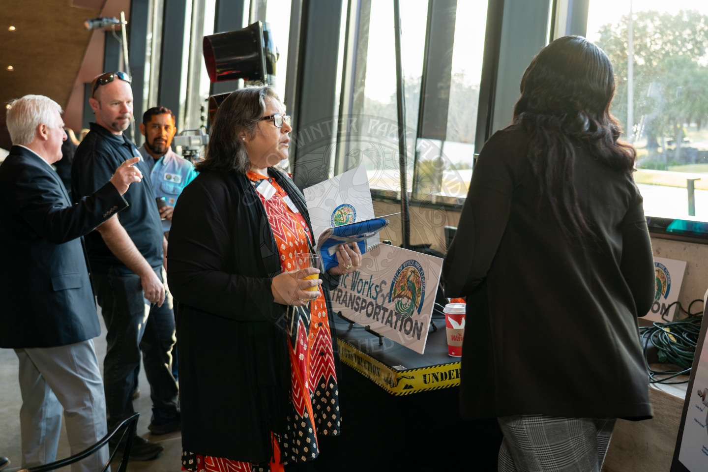 A few people stand and converse near a window at an indoor event. A woman in a red dress holds a blue plate, and event signs labeled “Public Works &amp; Transportation” are visible on a table beside her.