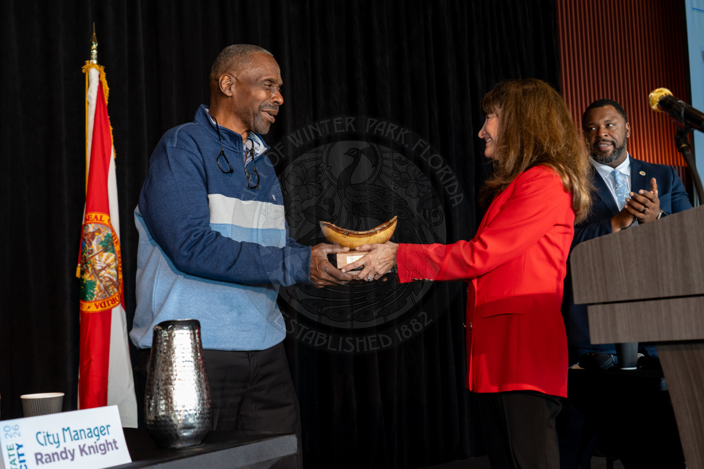 A woman in a red blazer hands a trophy to a smiling man in a blue sweater on stage, with another man applauding nearby.