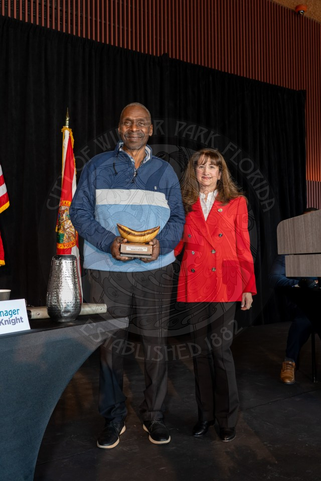 A man holding a trophy and a woman in a red jacket stand side by side on a stage, smiling at the camera.