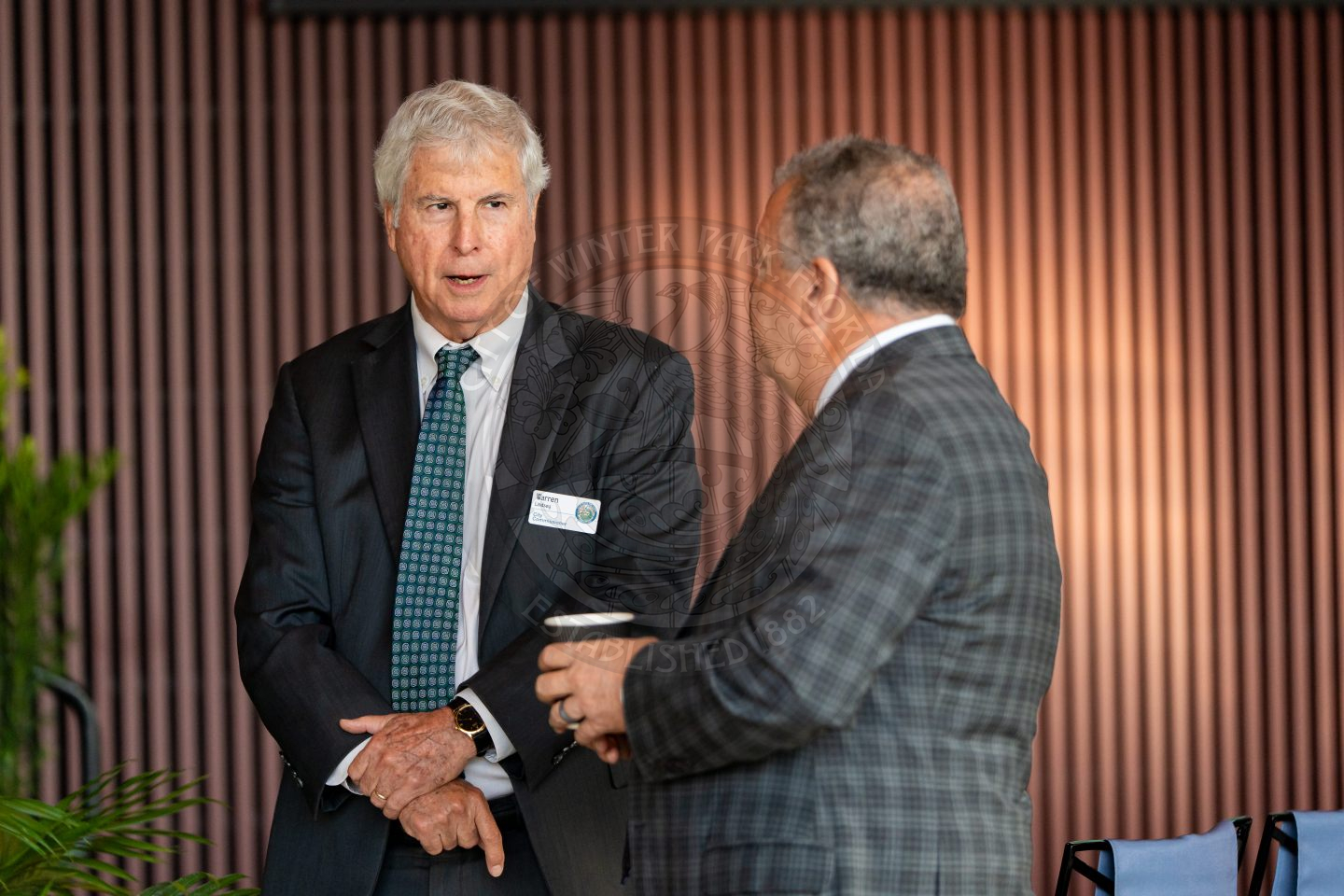 Two men in suits are having a conversation indoors while one is holding a coffee cup while the other speaks with his hands crossed.