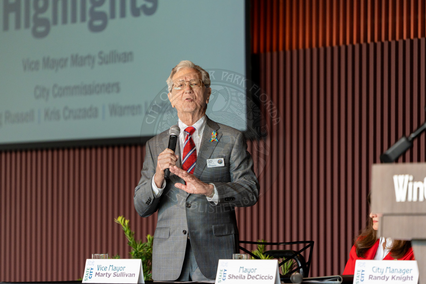 An elderly man in a suit and tie speaks into a microphone at a podium. A screen behind him displays text, and nameplates on the table in front of him read Vice Mayor Marty Sullivan and Mayor Sheila DeCiccio.