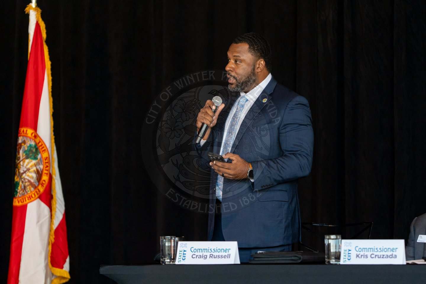 A man in a suit speaks into a microphone at a podium with the Florida state flag beside him. A nameplate reading Commissioner Craig Russell is on the table in front of him.