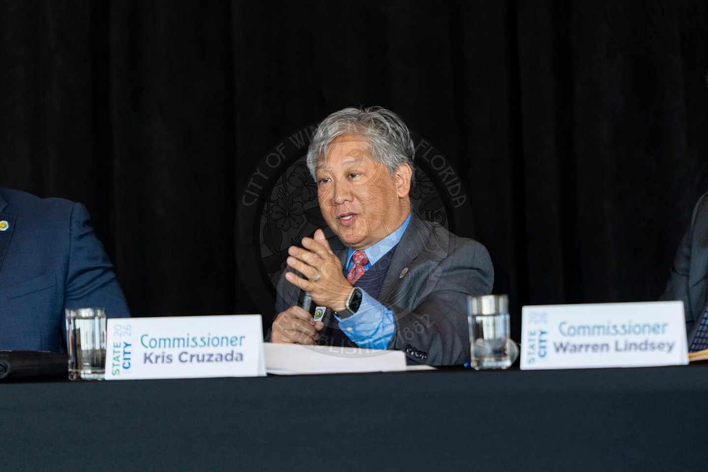 A man in a suit, identified as Commissioner Kris Cruzada by a nameplate, speaks while seated at a table with microphones, papers, and water glasses. A dark background and another nameplate are visible.